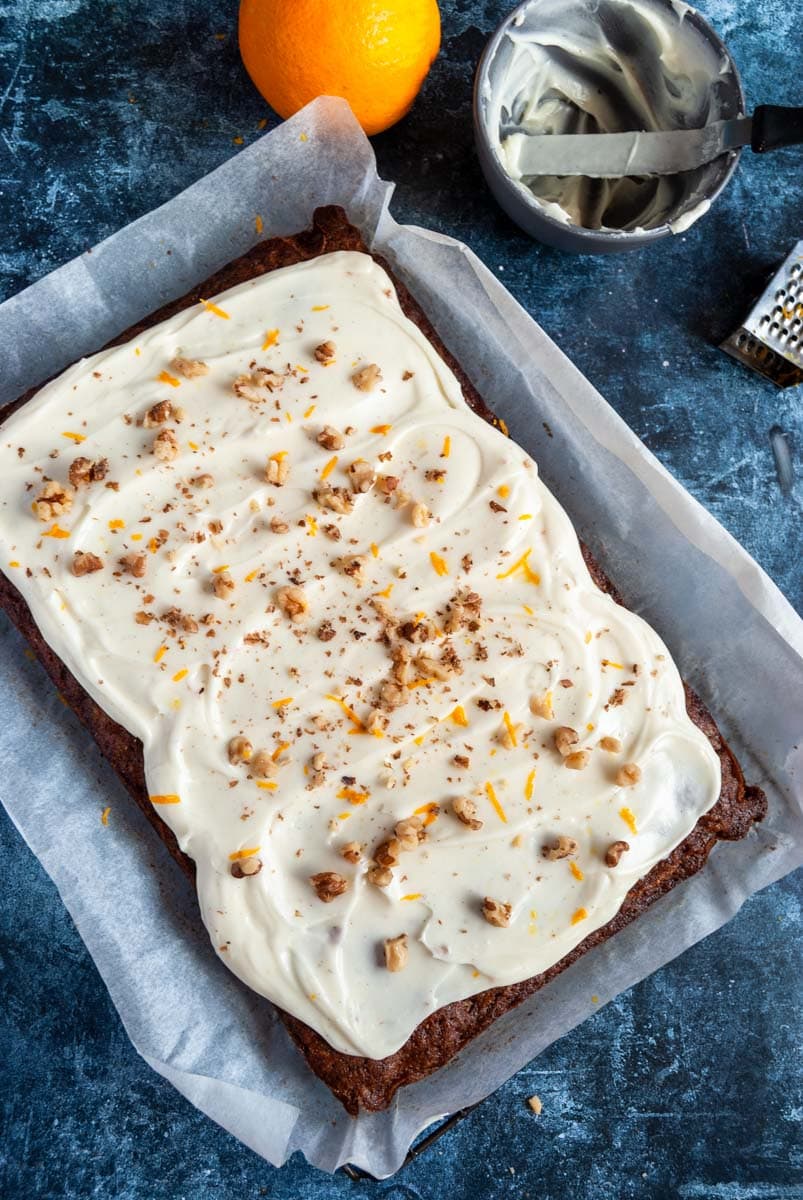 a large carrot cake with cream cheese icing sprinkled with chopped walnuts and orange zest, a small grey bowl with a small palette knife, a mini grater and an orange on a dark blue backdrop.