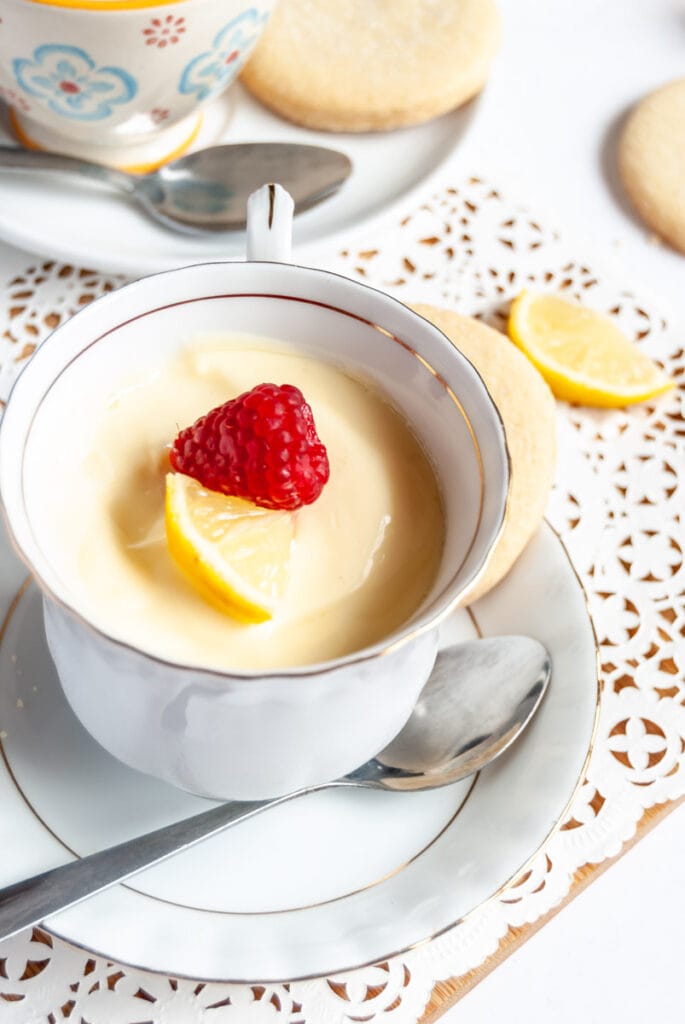 a white gold rimmed tea cup filled with lemon posset, topped with a fresh raspberry and lemon slice on a white gold rimmed saucer with a teaspoon.