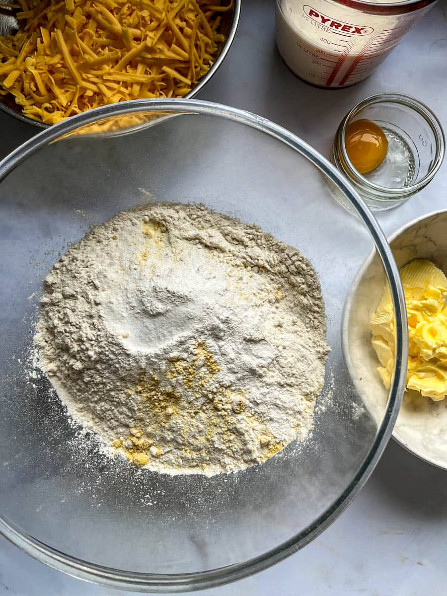 a glass mixing bowl of white flour and mustard powder, a white bowl of butter, a pot of egg yolk, a bowl of grated cheese and a jug of milk.