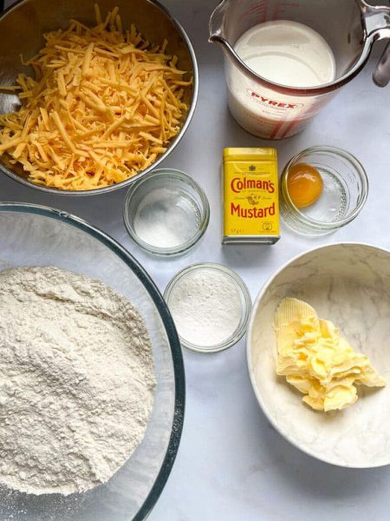 a glass mixing bowl of flour, a tin of dried mustard powder, a white bowl of butter, a jug of milk, a silver bowl of grated cheese, a small pot of egg yolk and two pots of baking powder and salt.