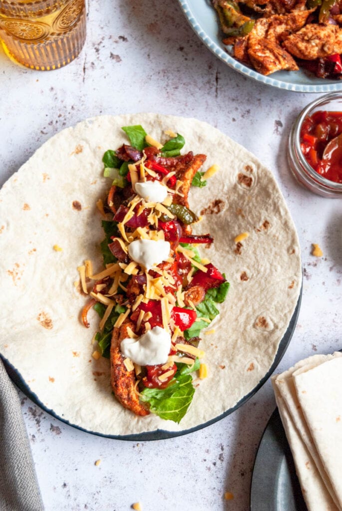 a chicken Fajita wrap with shredded lettuce, grated cheese, onions, bell peppers, grated cheese, salsa and sour cream on a black plate.