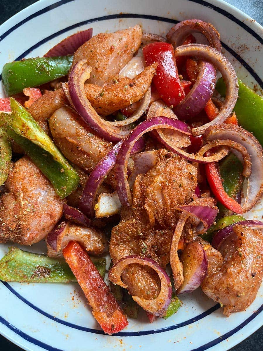 a white and blue striped bowl of raw chicken strips, sliced red onion and green and red bell peppers coated in a spice mix.