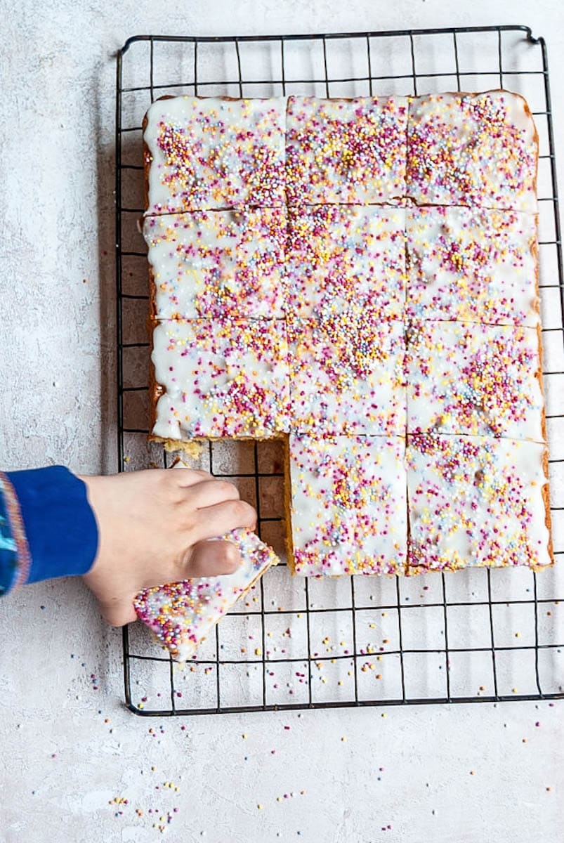 A vanilla sponge traybake covered in white icing and multi-coloured sprinkles cut into squares. A child's hand can be seen reaching for a slice.