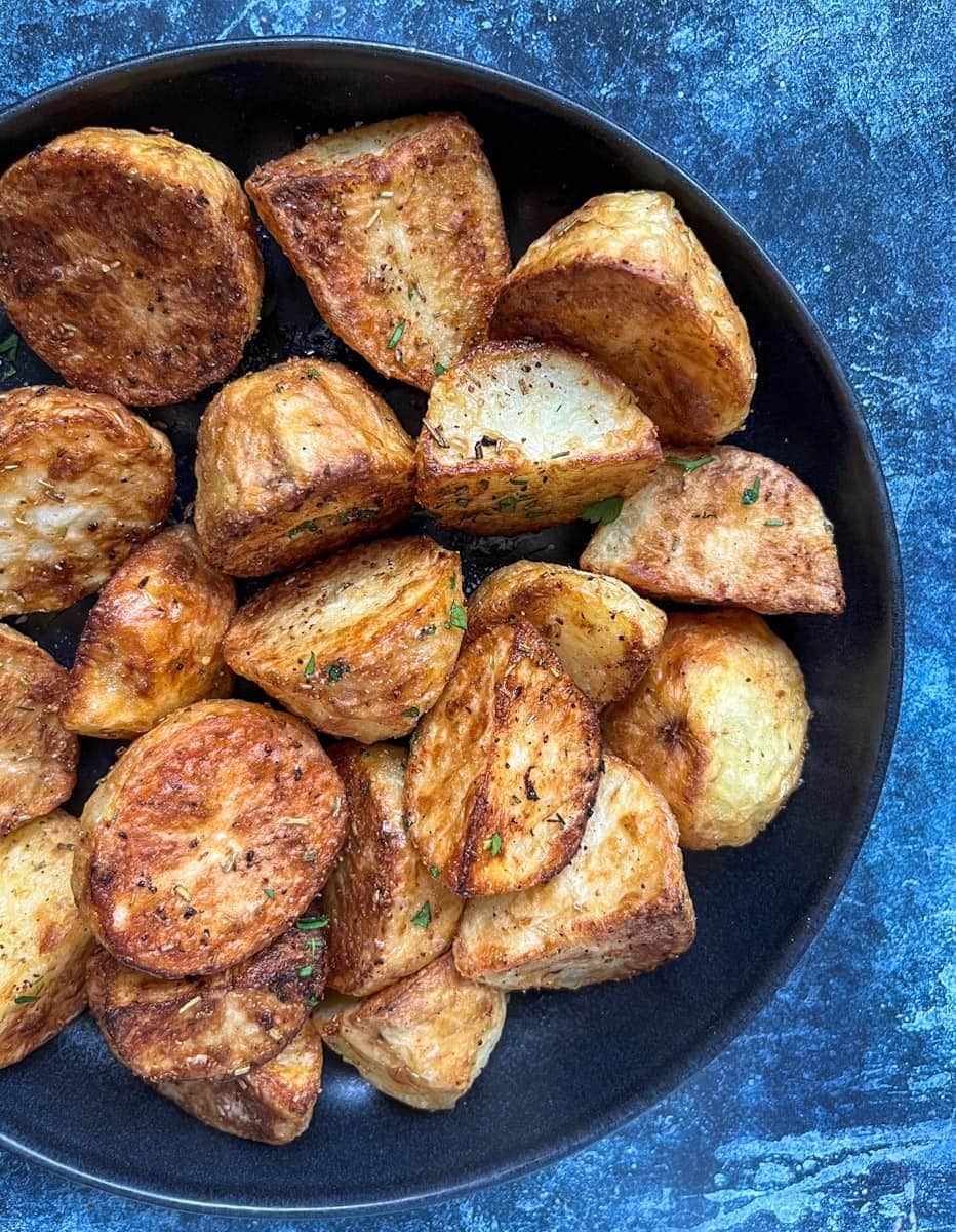 a black serving bowl of golden roasted potatoes on a blue backdrop.