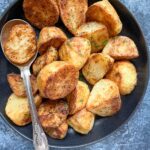 a black bowl of roasted potatoes with a large metal spoon on a blue backdrop.