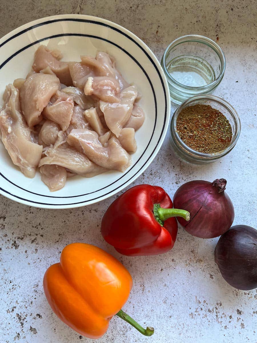 a blue and white bowl of raw chicken pieces, a yellow bell pepper, a red bell pepper, two red onions, a small pot of fajita spice mix and a pot of oil on a mottled grey and white backdrop.