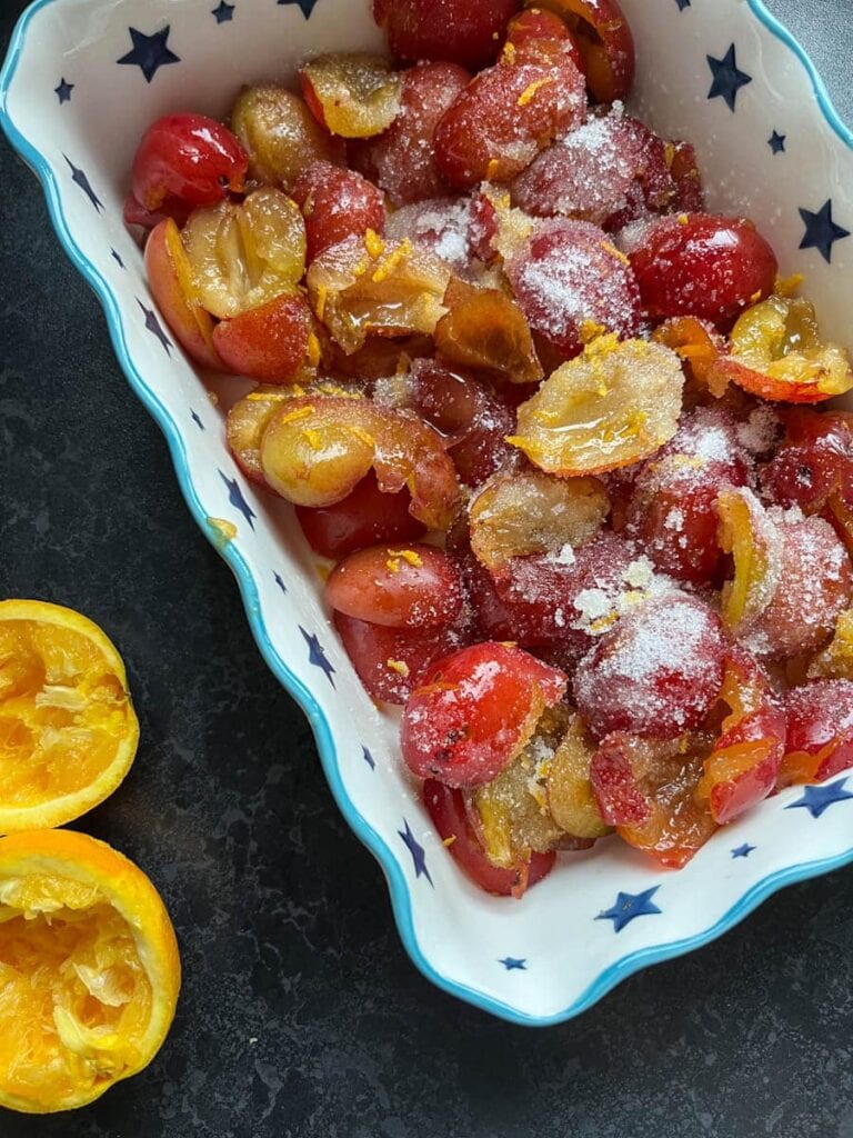 a white and blue star patterned baking dish of halved plums sprinkled with sugar and orange zest.