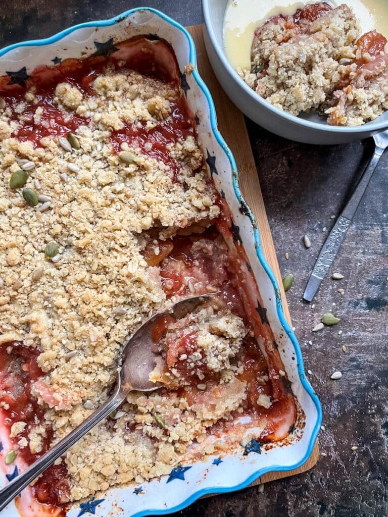 a white and blue star patterned dish of plum crumble with a large silver spoon and a bowl of plum crumble and custard.