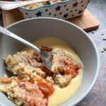 a grey bowl of plum crumble and custard with a silver spoon. A blue and white star patterened dish of crumble sits in the background.
