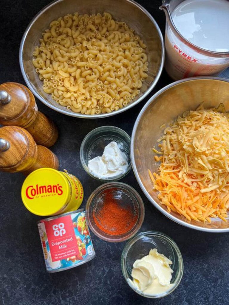 silver bowls of elbow macaroni, grated cheese, a jug of milk, wooden salt and pepper mills, small jars of cream cheese, butter and paprika, a jar of English mustard and a tin of evaporated milk.