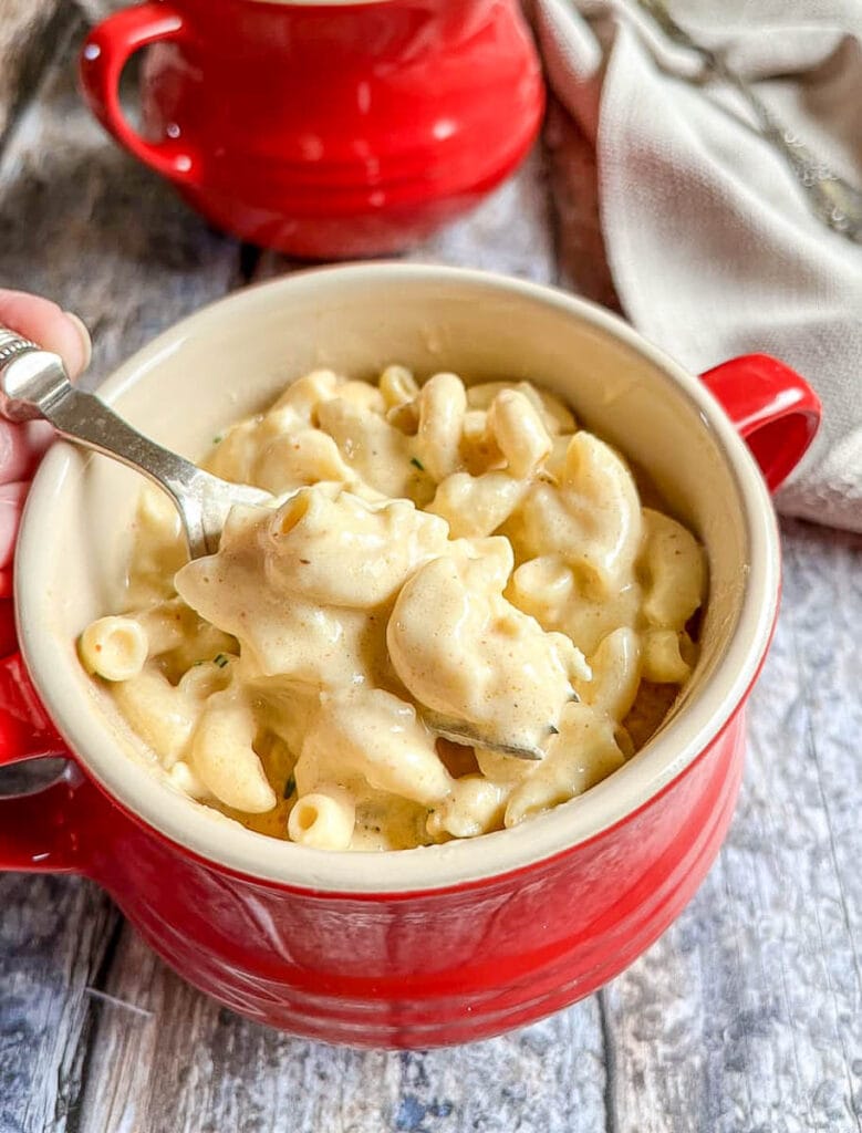 a red and cream bowl of macaroni cheese with a silver fork on a wooden table.