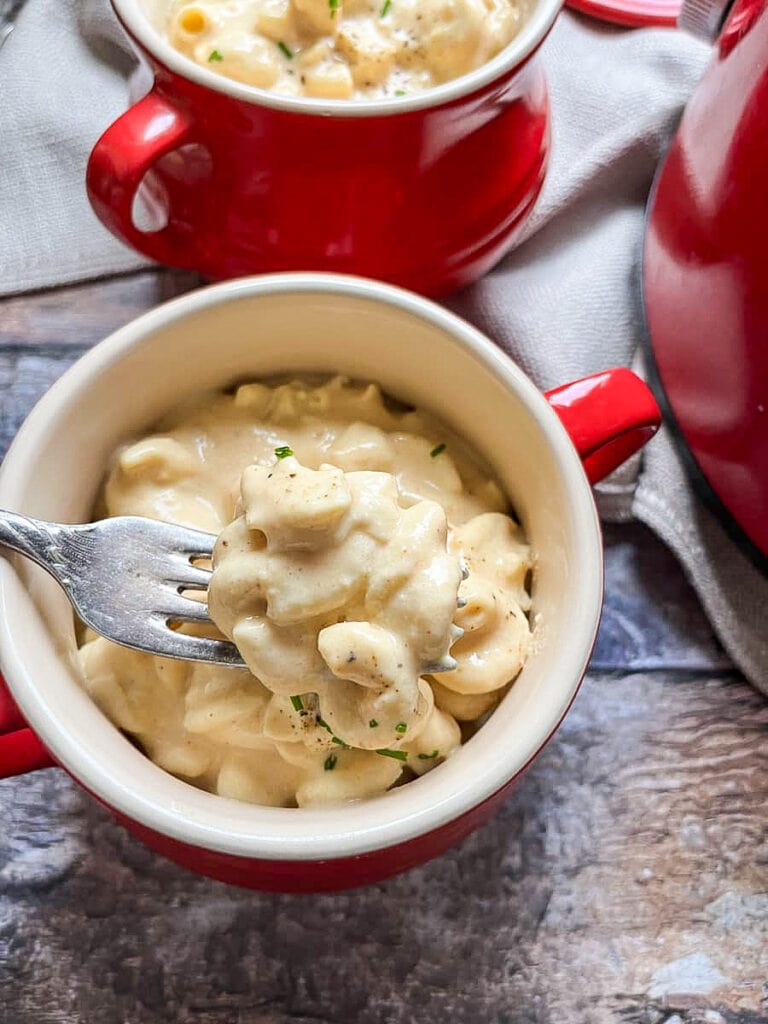 a red and cream bowl of macaroni cheese with a silver fork on a wooden table.