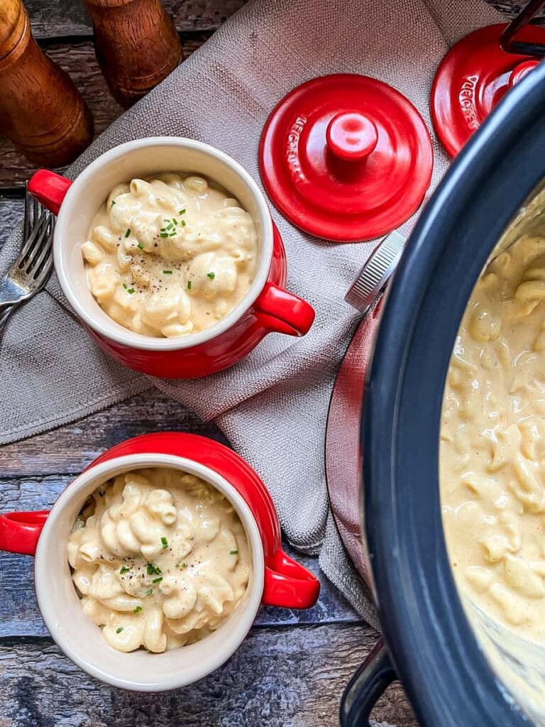 two red and cream bowls filled with macaroni cheese spinkled with fresh chives sitting on a wooden table with a grey napkin, wooden salt and pepper mills and a slow cooker filled with macaroni cheese.