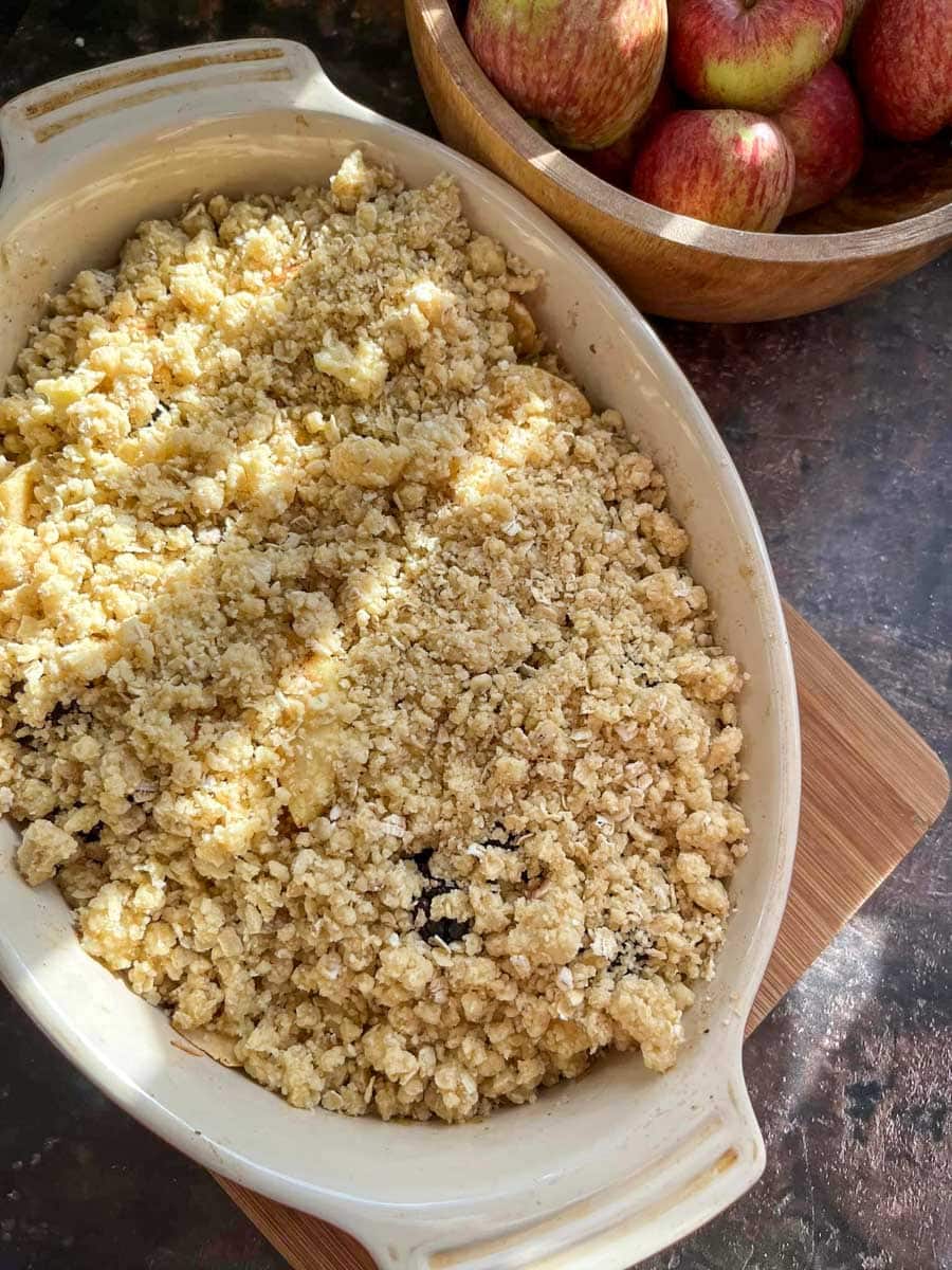 an apple crumble in a cream ceramic dish on a wooden board and a wooden bowl of apples.