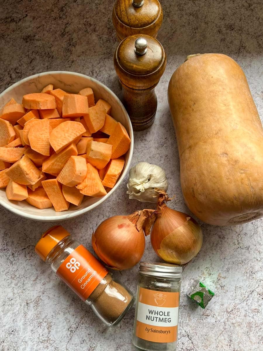a butternut squash, a white bowl of peeled and diced sweet potatoes, wooden salt and pepper mills, two onions, a bulb of garlic, a stock cube, a jar of ground cinnamon and a jar of whole nutmeg.