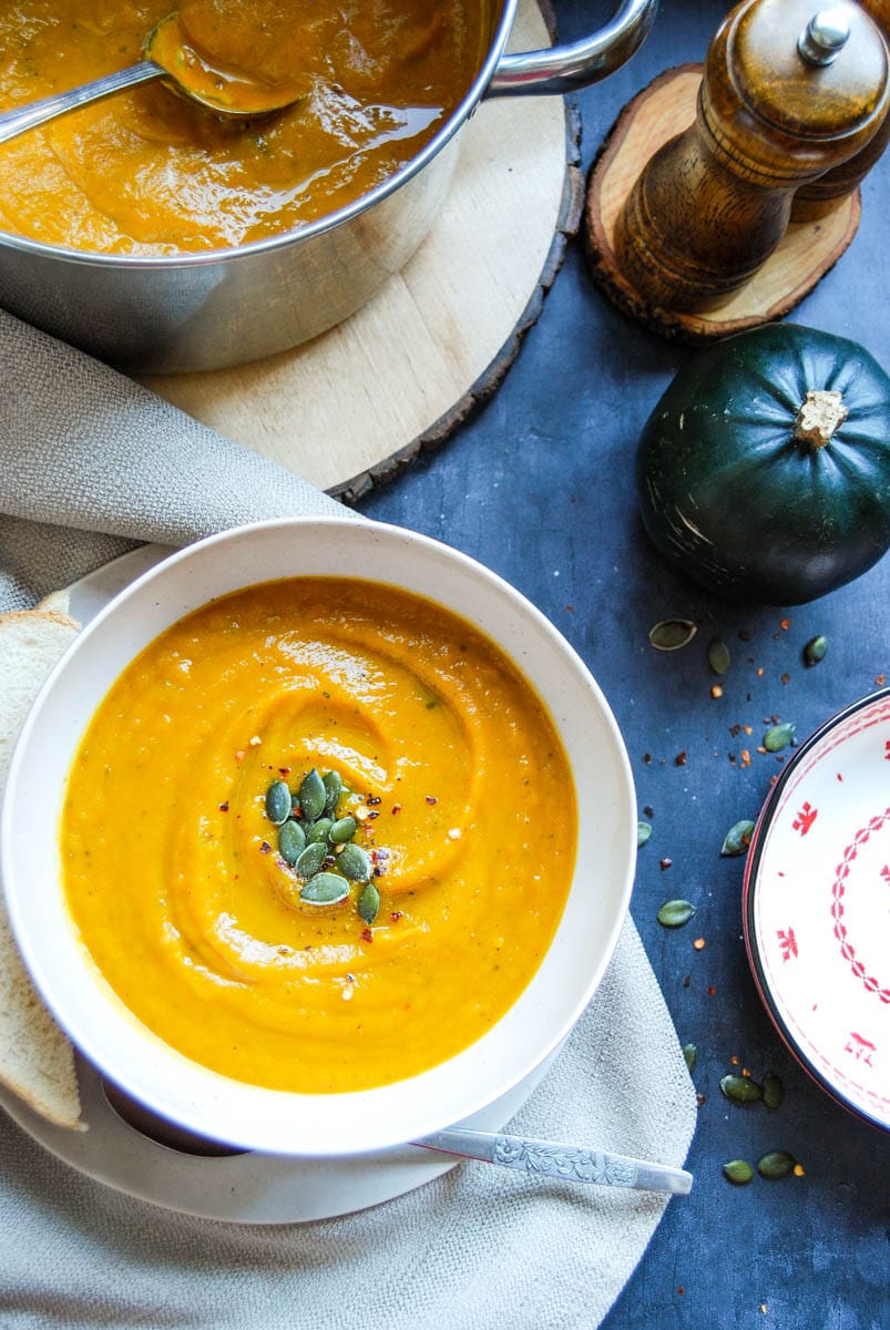 a bowl of pumpkin soup topped with red chilli flakes and pumpkin seeds, a beige linen napkin, a large saucepan of pumpkin soup, a small green pumpkin and wooden salt and pepper mills.