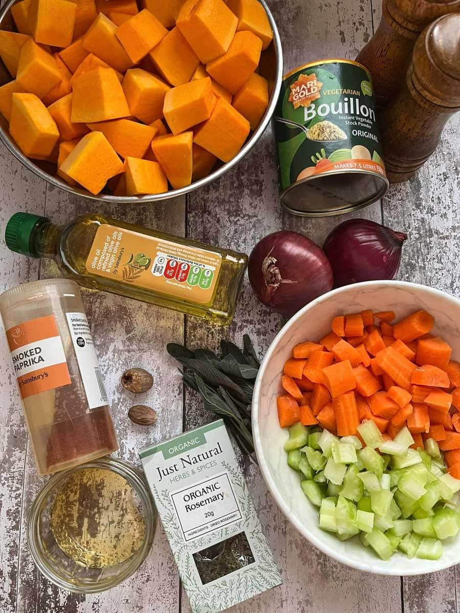 a silver bowl of diced pumpkin, a white bowl of chopped celery and carrots, two red onions, fresh sage leaves, nutmeg, a tub of smoked paprika, a bottle of olive oil, a tub of vegetable boullion and wooden salt and pepper mills.