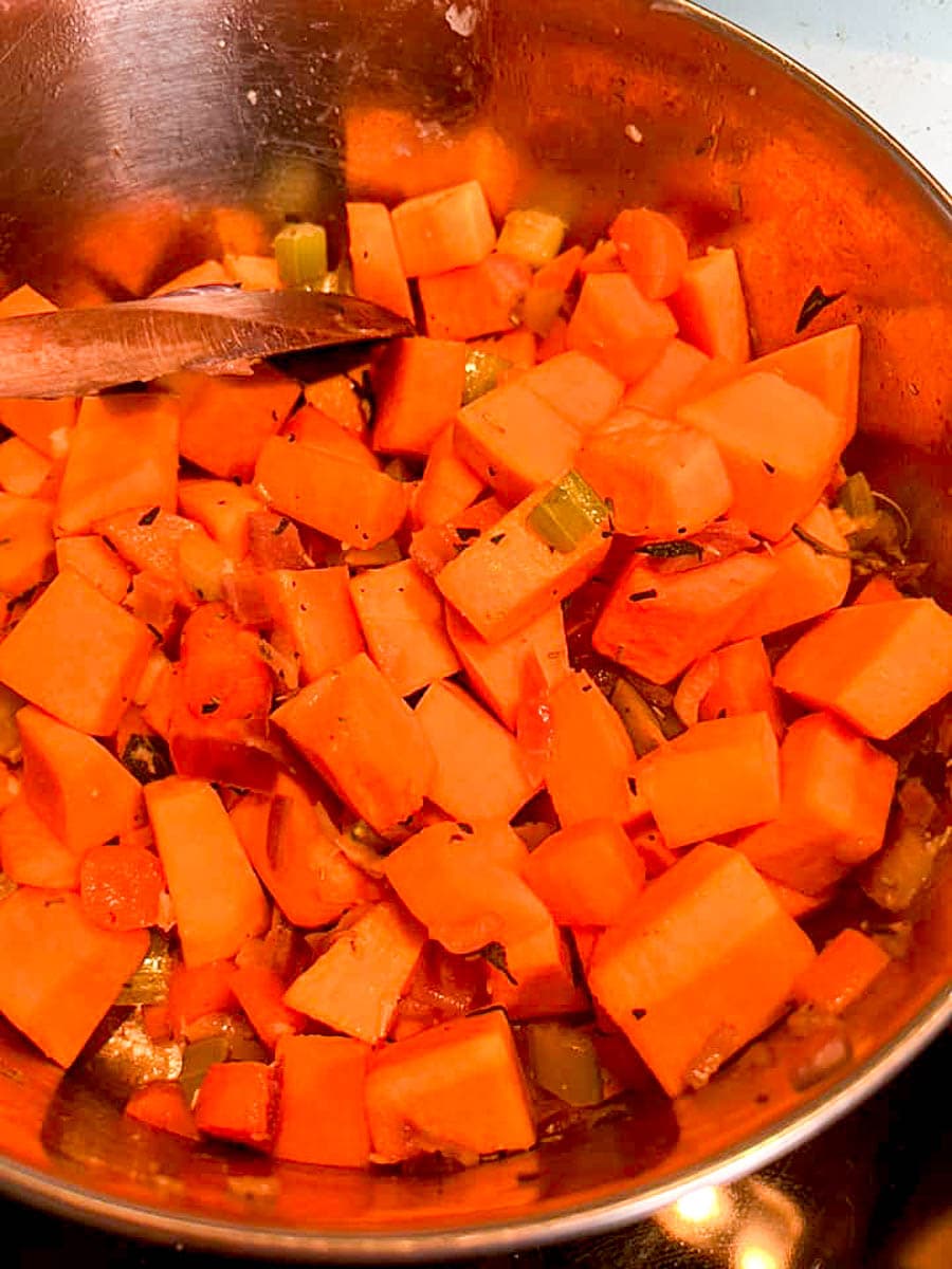 diced pumpkin, onion, celery, carrot and herbs cooking in a silver pan with a wooden spoon.