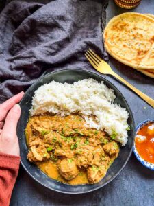 a black stoneware bowl of chicken curry and rice, a blue pot of mango chutney, naan bread and a black linen napkin.