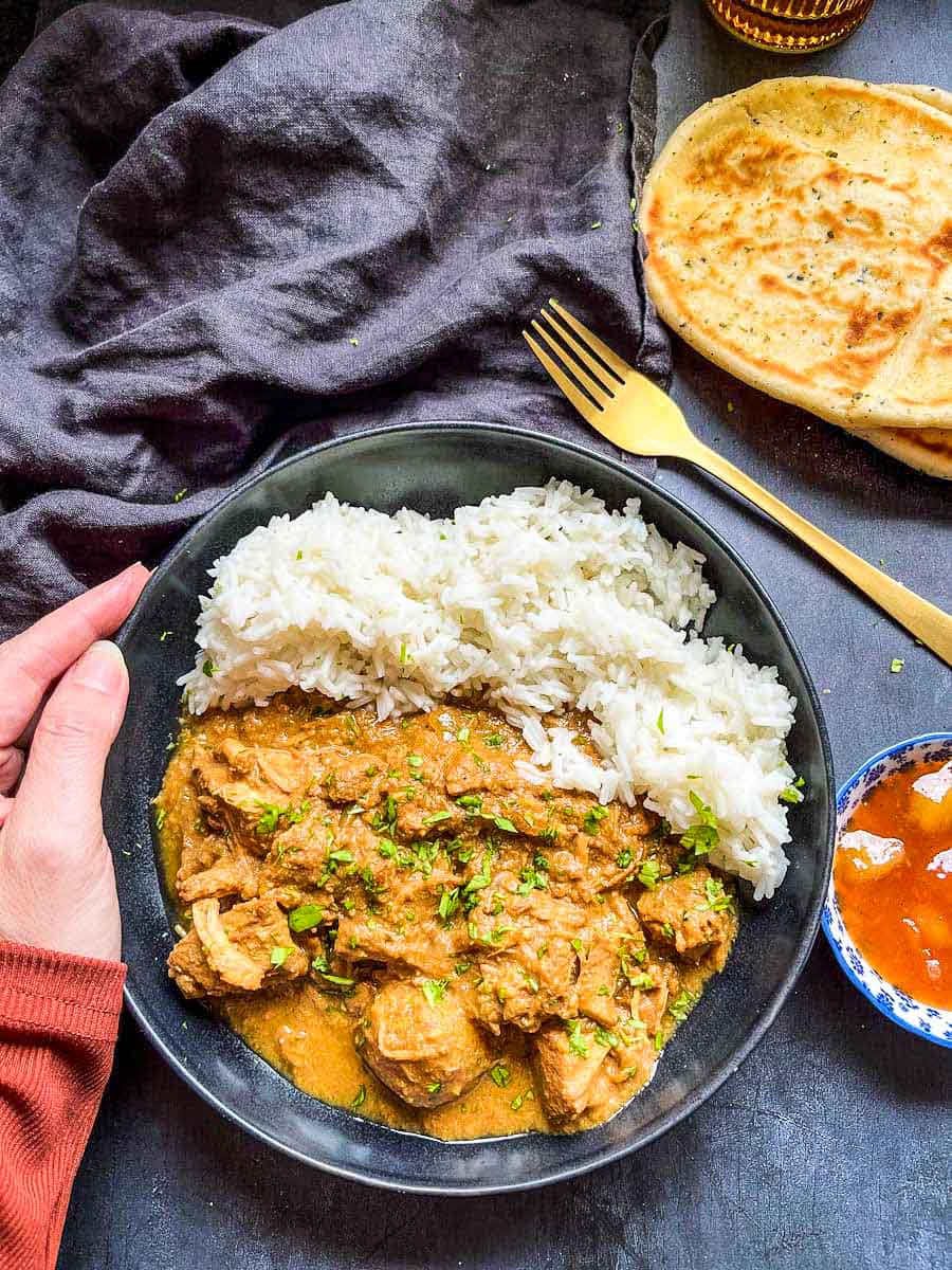 a black stoneware bowl of chicken curry and rice, a blue pot of mango chutney, naan bread and a black linen napkin.