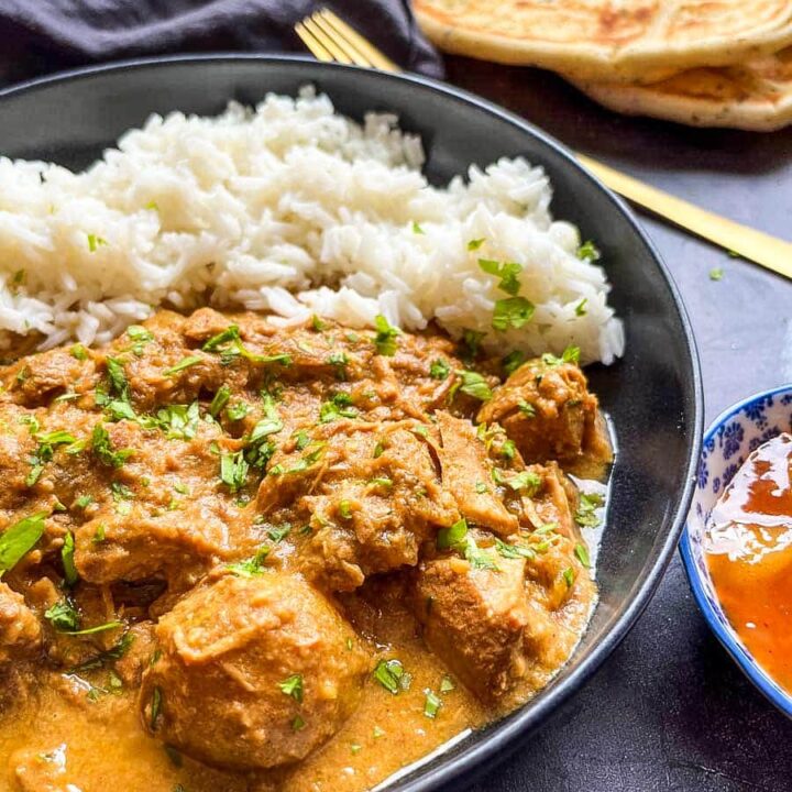 a black stoneware bowl of chicken curry with rice. A gold fork, a black napkin and naan bread can be seen in the background.