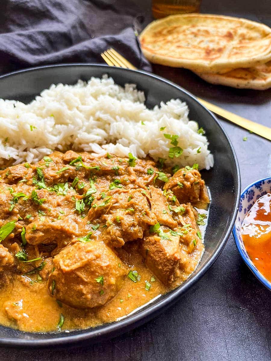 a black stoneware bowl of chicken curry with rice. A gold fork, a black napkin and naan bread can be seen in the background.