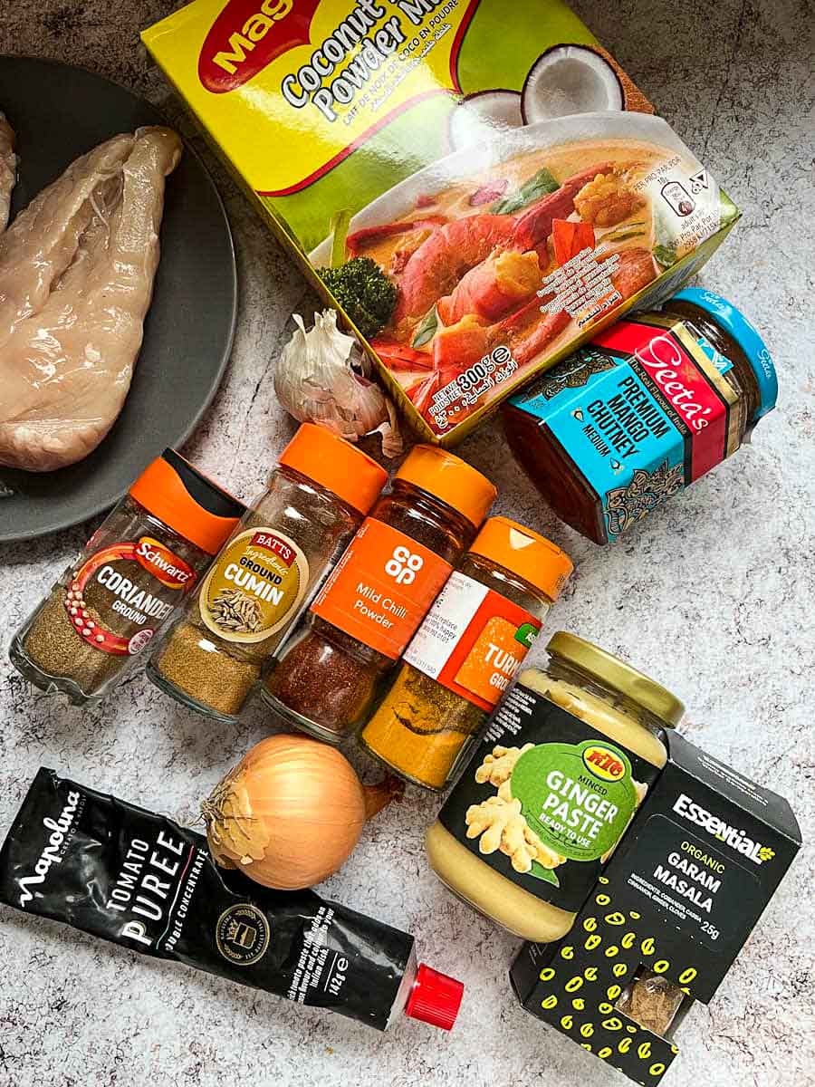 a black plate of boneless chicken fillets, a box of coconut milk powder, a jar of mango chutney, an onion and a bulb of garlic, a jar of ginger paste, tomato puree, and jars of spices.