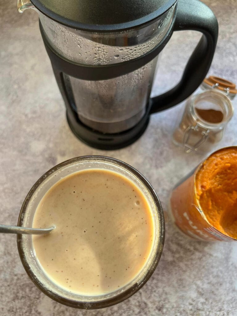 A cafetière filled with coffee, a can of pumpkin purée, a small jar of pumpkin spice and a latte glass filled with coffee and spiced milk.