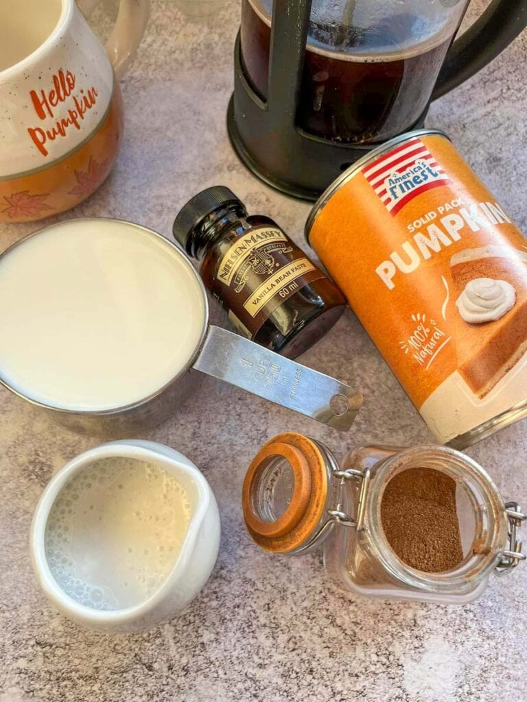 A silver cup of milk, a tin of pumpkin purée, a jar of spice, a bottle of vanilla extract, a coffee pot and a pumpkin mug.