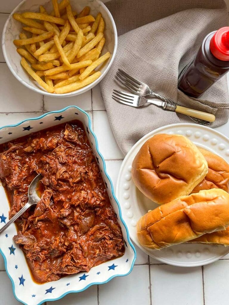 a blue and white star patterned dish of shredded chicken in bbq sauce with a silver spoon, a white plate of brioche rolls, a white bowl of fries and a bottle of bbq sauce with two forks and a beige linen napkin.