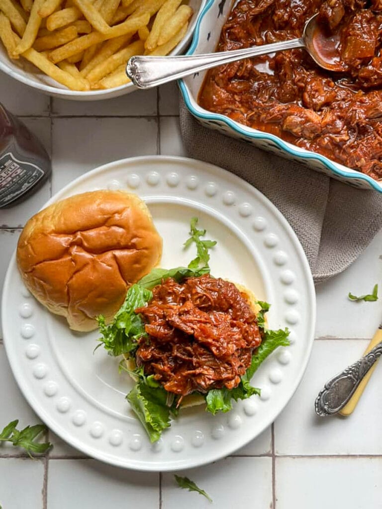 a brioche bun topped with lettce and shredded chicken in BBQ sauce on a white plate with a bowl of fries.
