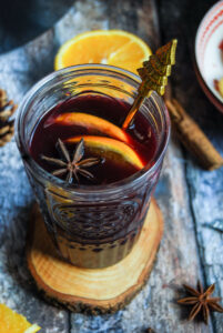 a close up photo of a glass of mulled wine sitting on a wooden coaster, with orange slices, star anise and a golden Christmas tree spoon.