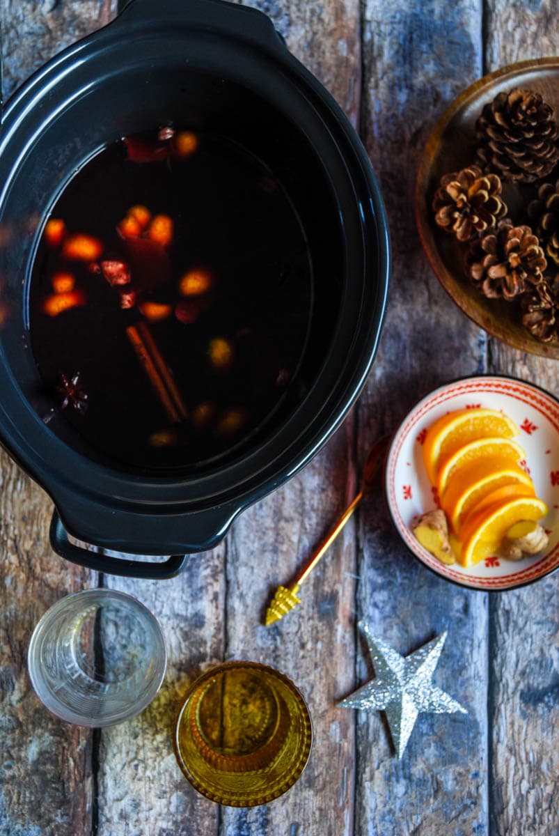 a slow cooker filled with mulled wine with cinnamon sticks, orange peel and star anise, a red and white bowl of oranges, a wooden bowl or pine cones, a silver star and two decorative glasses on a faded wooden background.