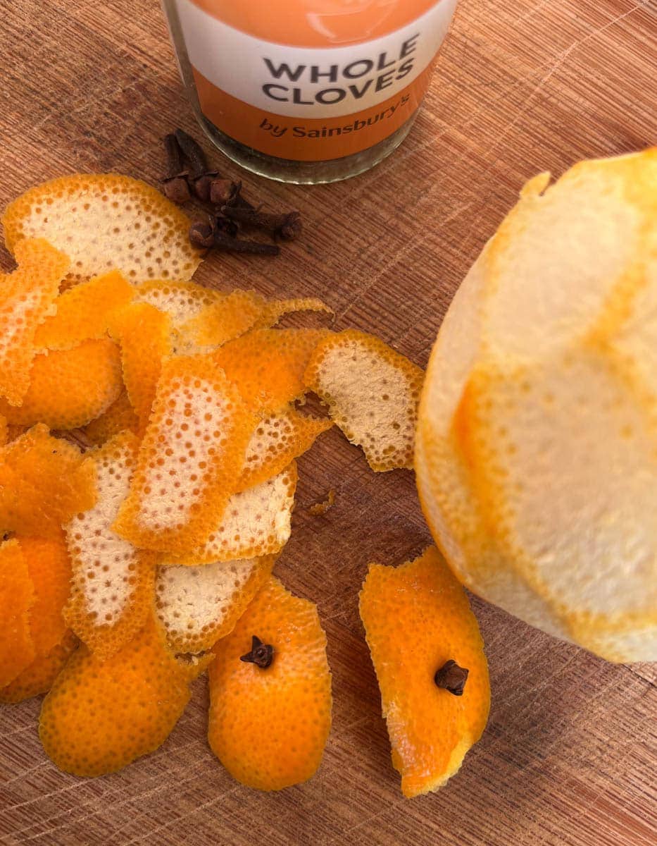 orange peel, whole cloves and a peeled orange on a wooden chopping board.