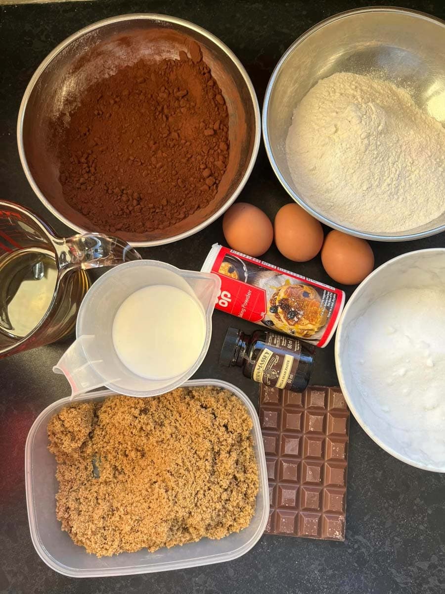 various bowls of cocoa powder, flour and sugar, jugs of milk and oil, three eggs, a tub of brown sugar, baking powder, vanilla extract and a bar of chocolate.