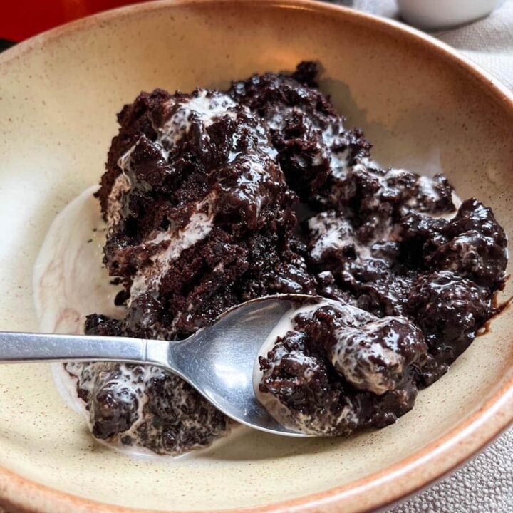 a beige stoneware bowl of chocolate fudge puding and cream with a silver spoon. A red crock pot and a small white jug can be partially seen in the background.