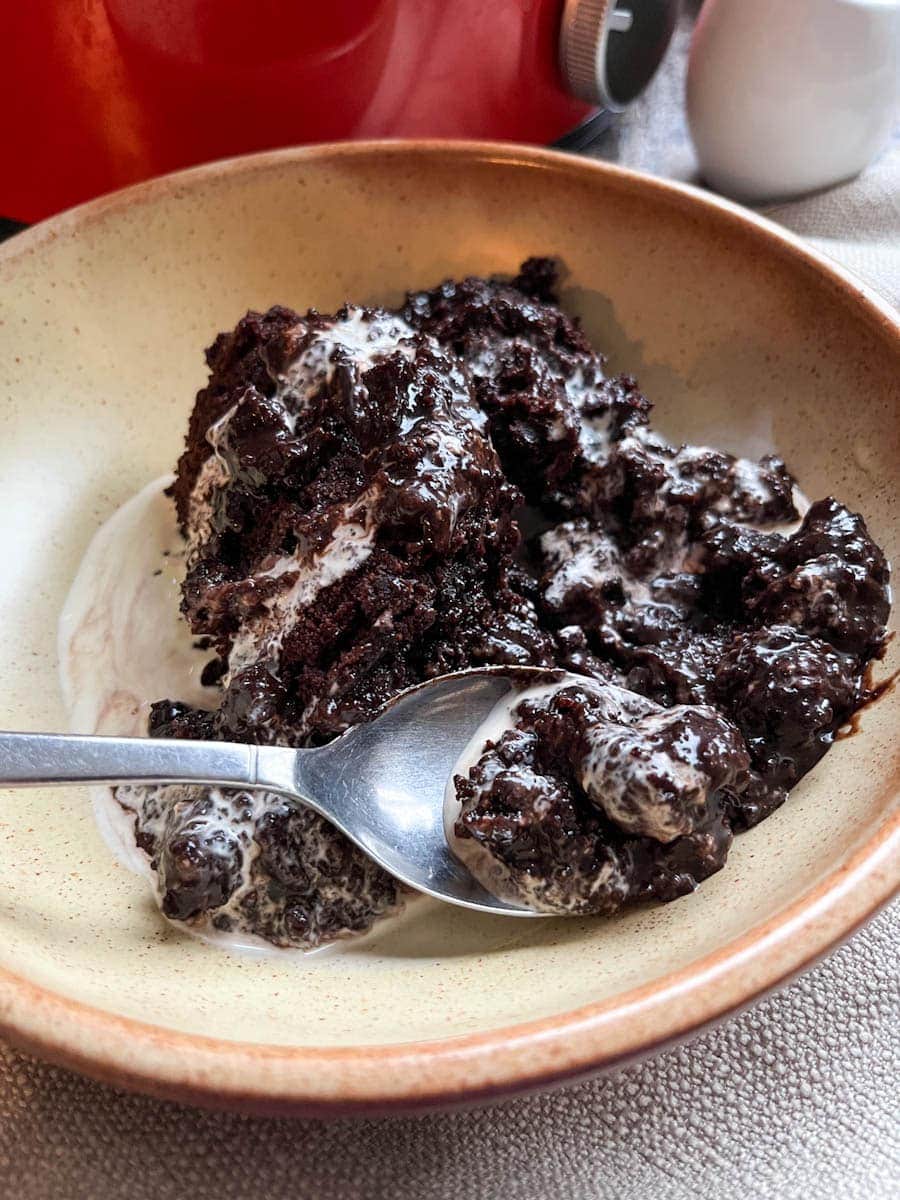 a beige stoneware bowl of chocolate fudge puding and cream with a silver spoon. A red crock pot and a small white jug can be partially seen in the background.