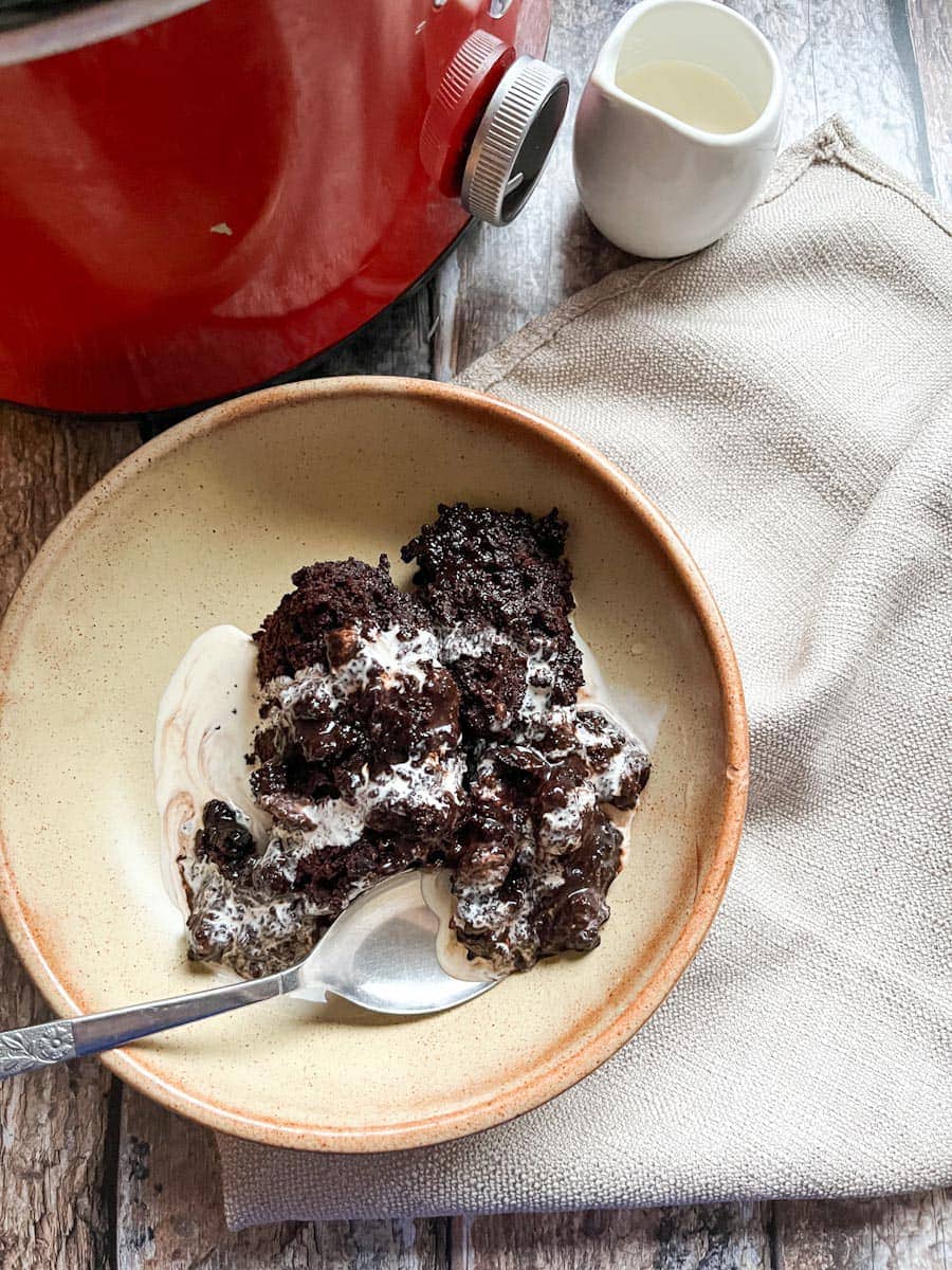 a beige stoneware bowl of chocolate fudge pudding, cream and a silver spoon, a grey napkin, a small pot of cream and a red crock pot.