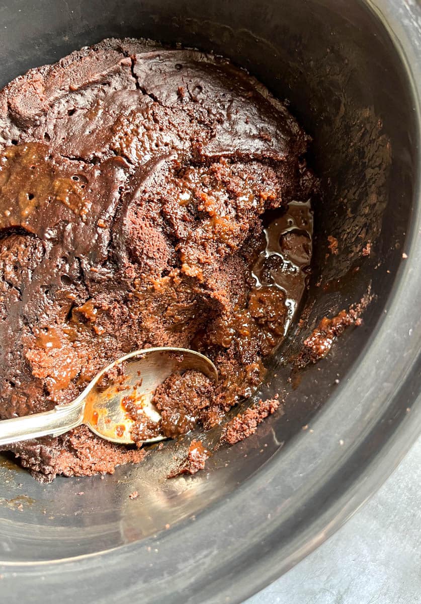 a sticky chocolate pudding cake in a grey slow cooker with a large silver spoon.