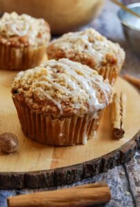 three large apple muffins topped with crumble and a vanilla glaze on a wooden board with cinnamon sticks.