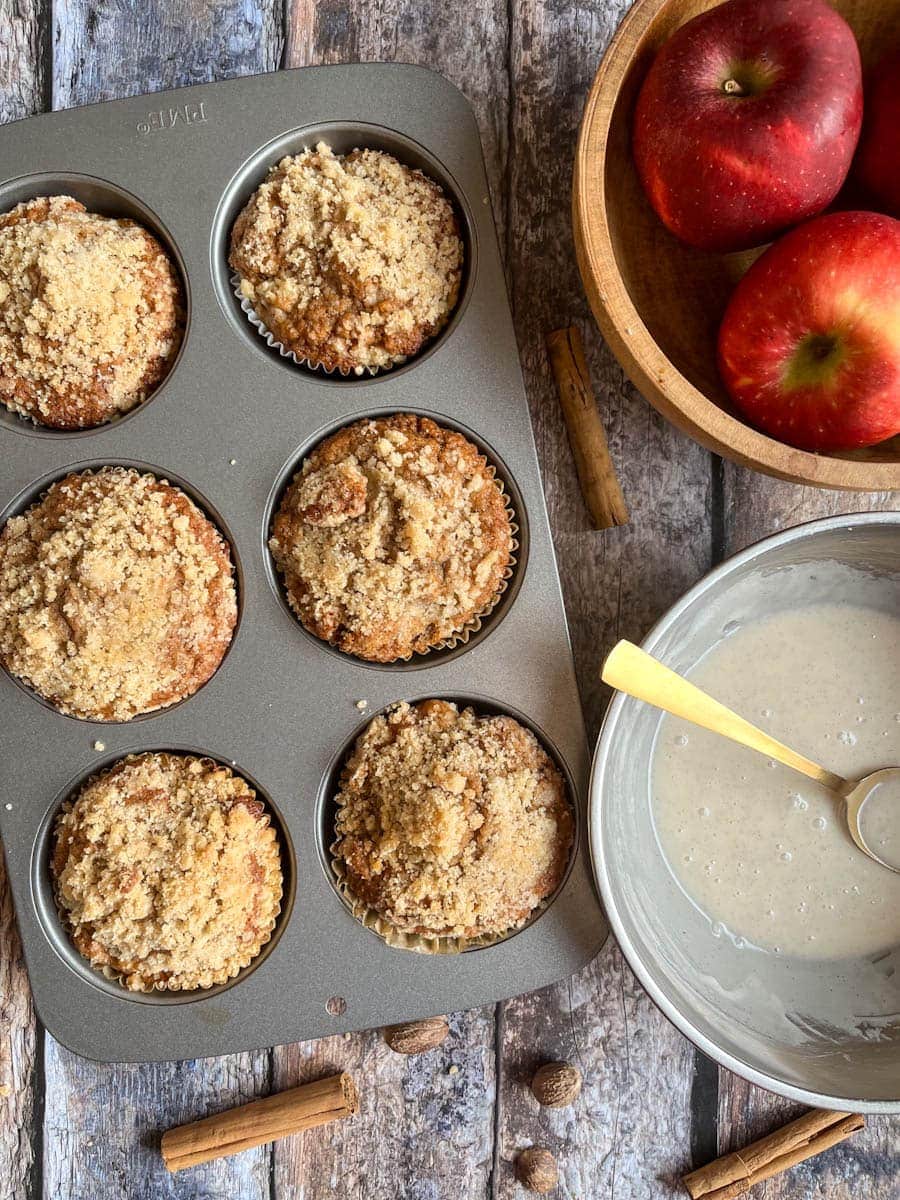 six large apple muffins in a jumbo muffin pan, a silver bowl of vanilla icing and a bowl of red apples on a wooden backdrop.