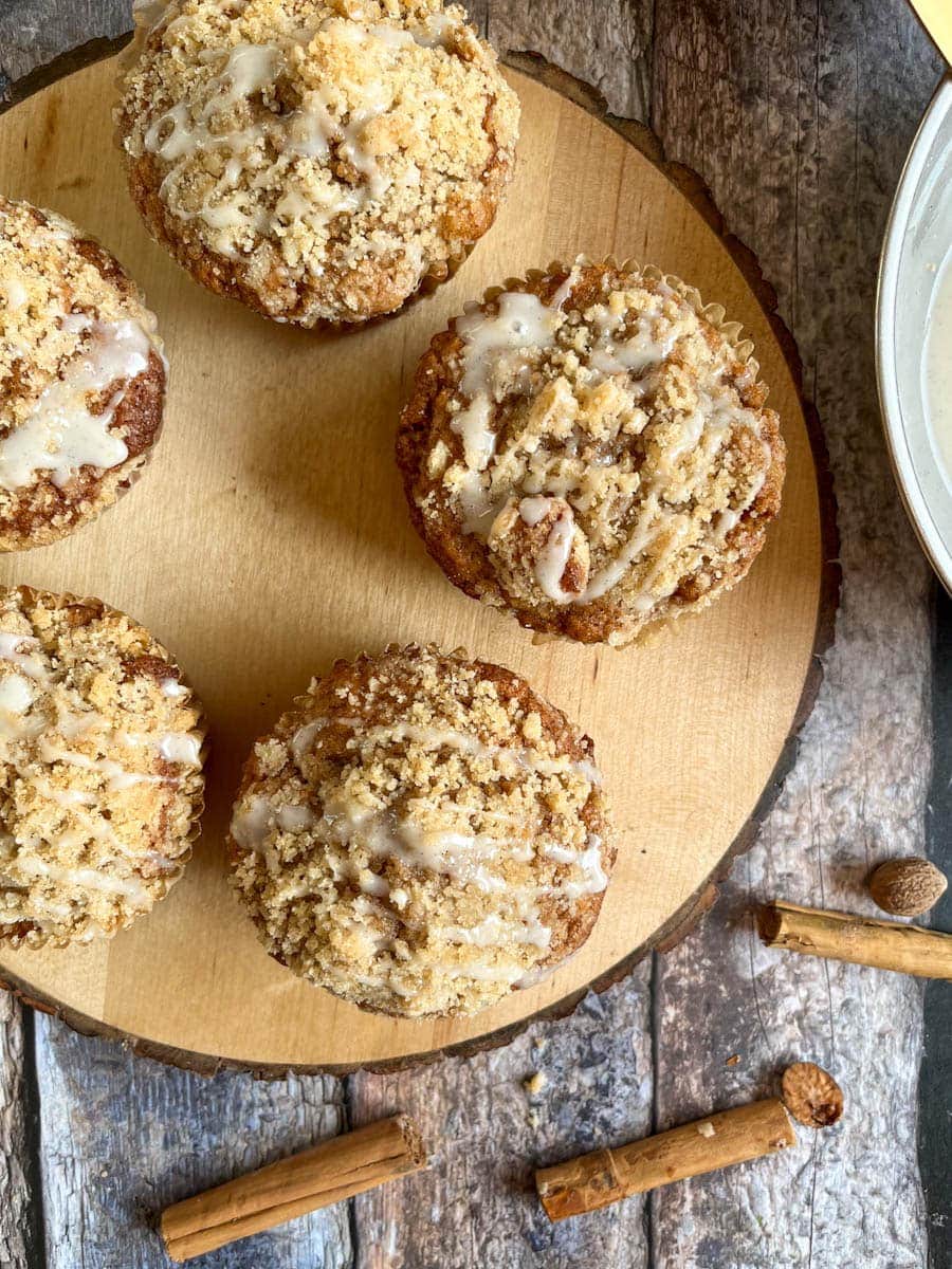 five large apple muffins topped with crumble and a vanilla glaze icing on a wooden board.