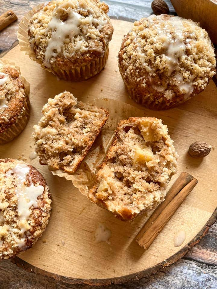 five apple muffins topped with crumble and vanilla glaze icing on a round wooden board.