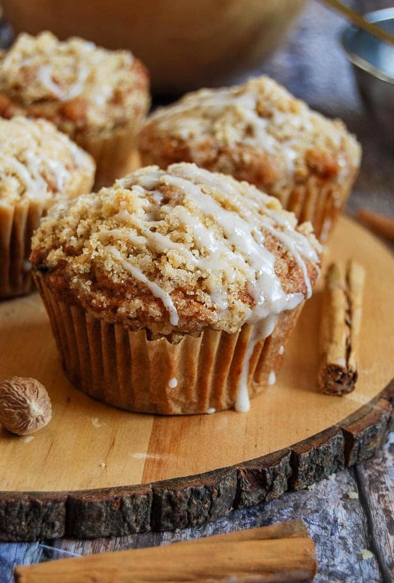 three large apple muffins topped with crumble and a vanilla glaze on a wooden board with cinnamon sticks.