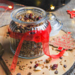A glass jar of granola with dried cranberries and nuts. A red ribbon is tied around the jar. The jar sits on top of a wooden serving board, with Christmas decorations and fairy lights.