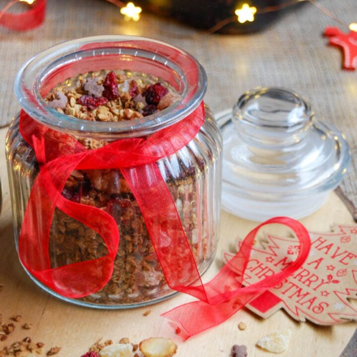 A jar of granola with cranberries and mixed nuts with a red ribbon tied around the jar sitting on top of a wooden serving board.