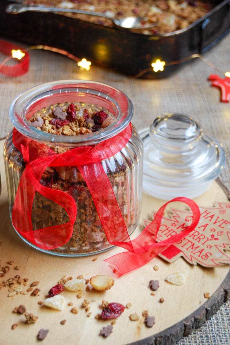 A jar of granola with cranberries and mixed nuts with a red ribbon tied around the jar sitting on top of a wooden serving board.