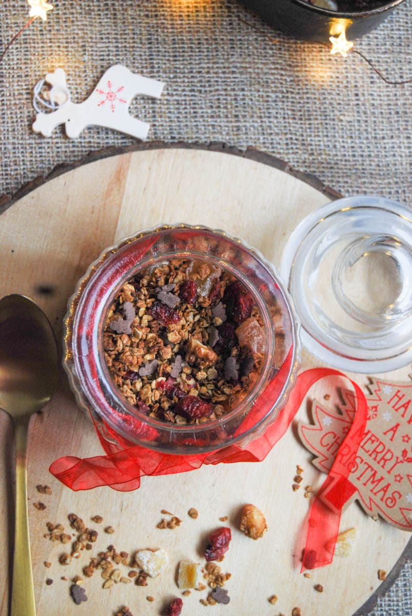 A flatlay photo of a jar of granola with cranberries and nuts tied with a red ribbon on a wooden serving board.