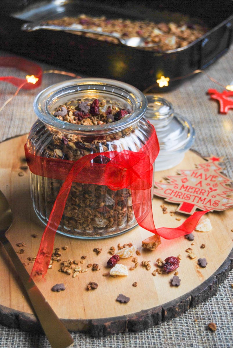 A glass jar of granola with dried cranberries and nuts. A red ribbon is tied around the jar. The jar sits on top of a wooden serving board, with Christmas decorations and fairy lights.