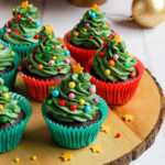 Chocolate cupcakes decorated with green buttercream icing, coloured round sprinkles and a gold star to resemble a Christmas tree on a wooden serving board.